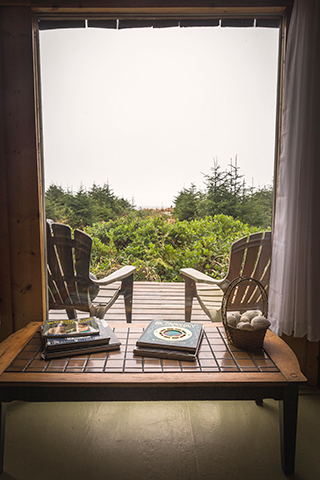 View from a cabin doorway: two deck chairs, a low table with books and a basket of shells, dune greenery beyond